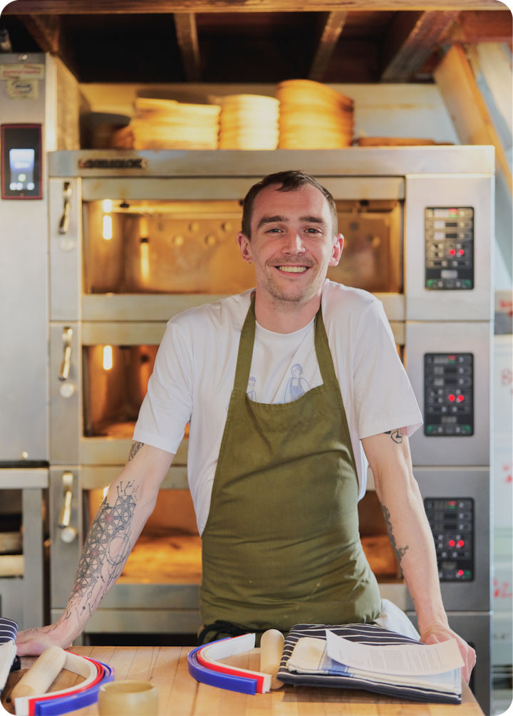 Une personne portant un tablier vert se tient à un comptoir en bois devant un four professionnel. Des ustensiles de pâtisserie et du papier sont soigneusement disposés sur le comptoir.