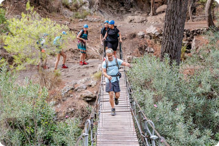Un groupe de randonneurs portant des casques et des sacs à dos explorent une zone boisée. Une personne traverse un étroit pont en bois entouré de verdure.