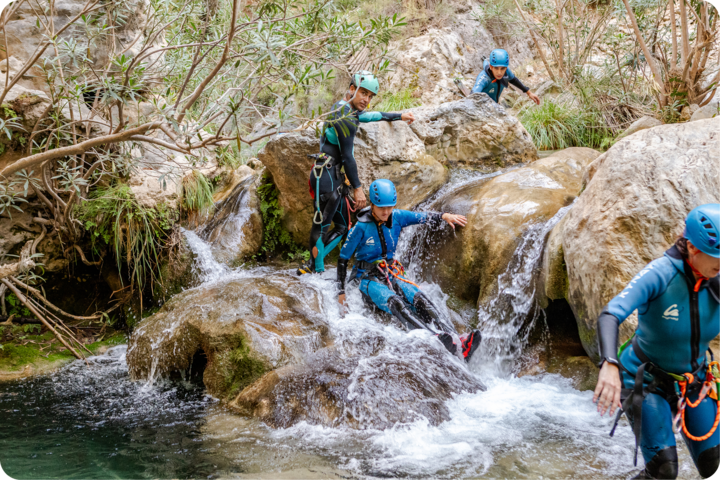 Des participants à une activité de plein air traversent un ruisseau en cascade sur un terrain rocheux accidenté, entourés de verdure et d'arbres.