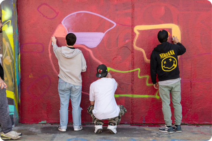 Un groupe de trois personnes crée des graffitis colorés sur un mur rouge, en utilisant de la peinture à la bombe pour ajouter des formes abstraites.