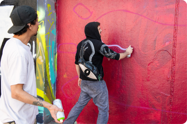 Une personne peint des lignes abstraites à la bombe sur un mur rouge, tandis qu'une autre tient des bombes de peinture, entourée de graffitis colorés.