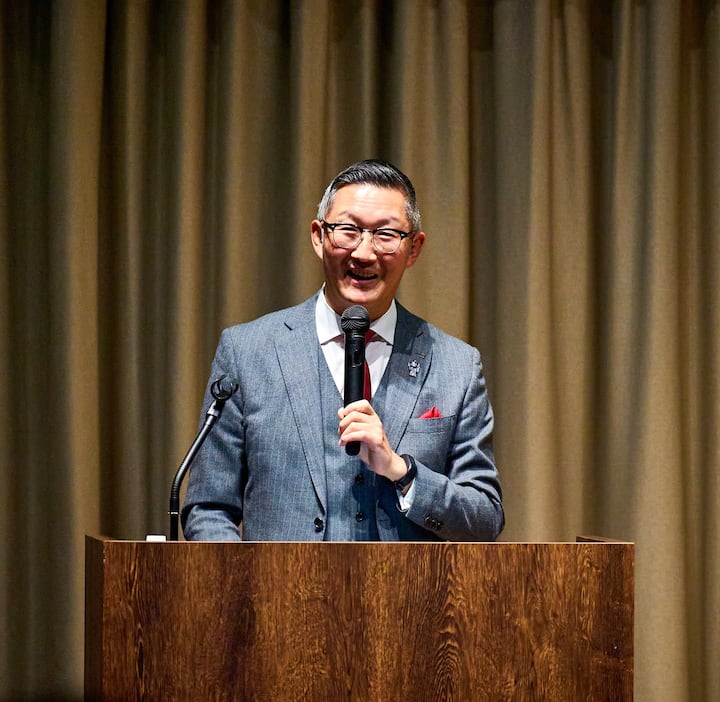 A person in a formal gray suit with a red pocket square speaks into a microphone while standing at a wooden podium in front of beige curtains.