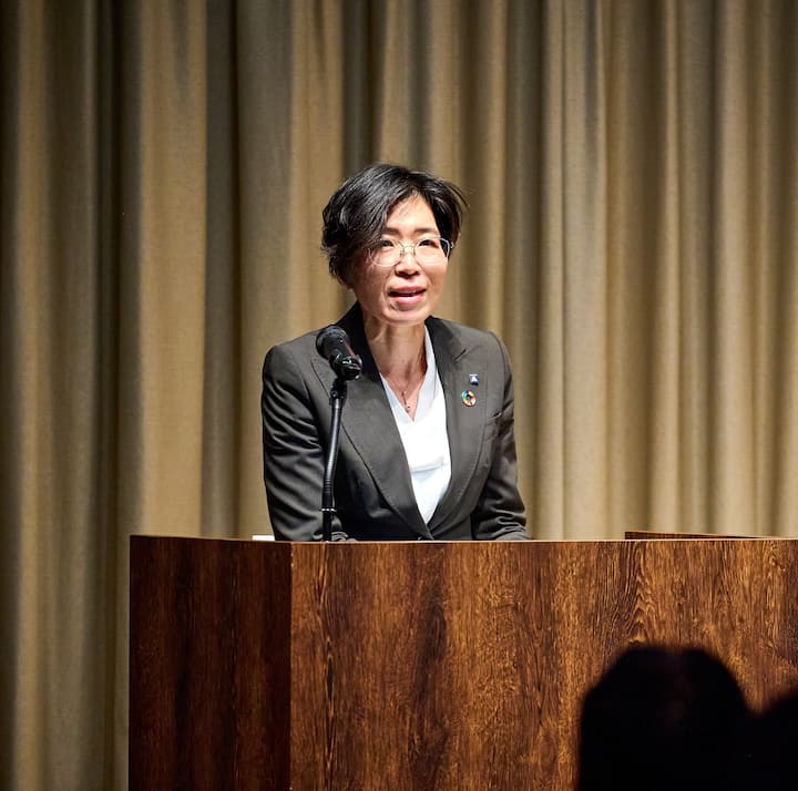 A person standing at a wooden podium with a microphone, wearing a formal suit and a pin displaying the Sustainable Development Goals emblem, with beige curtains in the background.