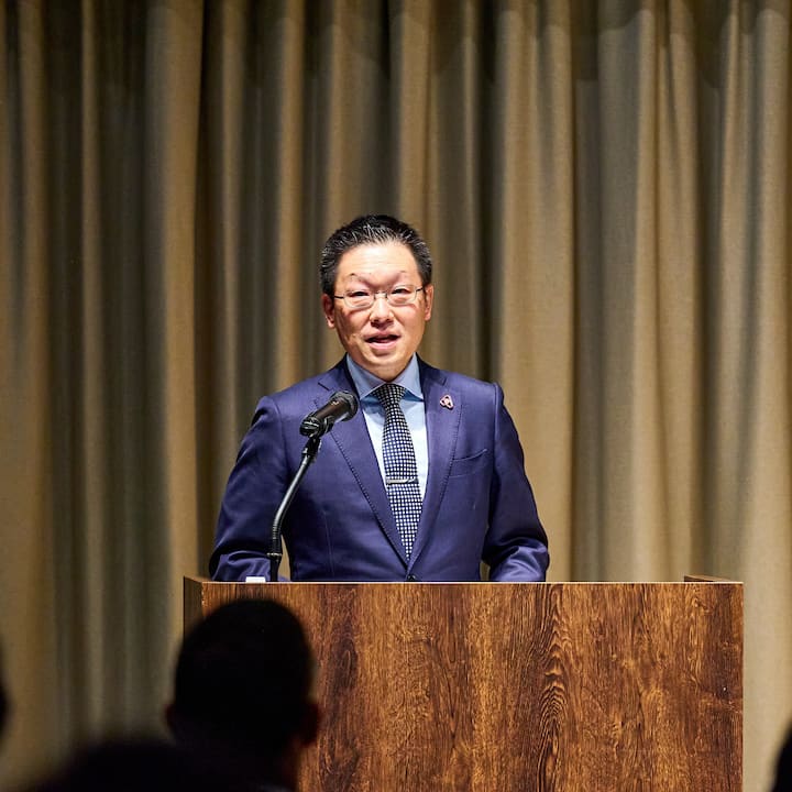 A speaker in a navy suit standing at a wooden podium, speaking into a microphone, with beige curtains in the background.
