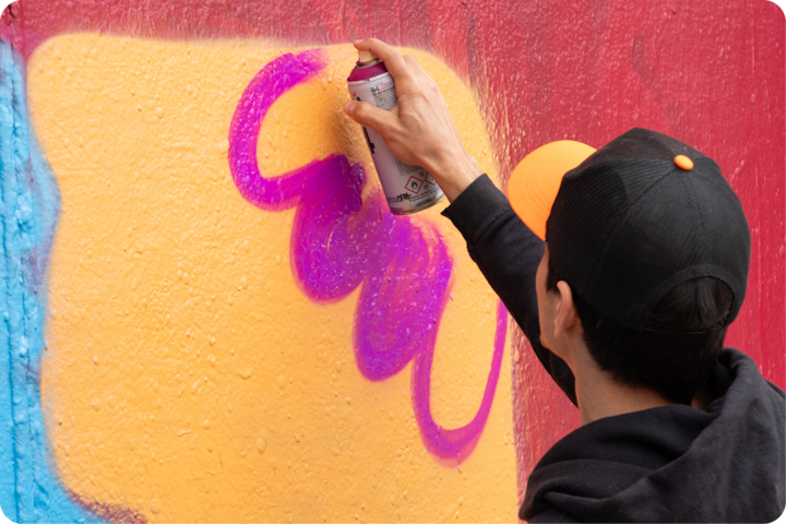 Une personne peint des formes violettes à la bombe sur un mur jaune et rouge vif dans le cadre d'une activité artistique.