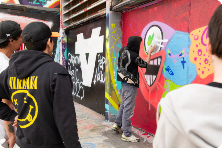 Une personne crée une fresque à la bombe sur un mur rouge, utilisant des motifs lumineux et ludiques, sous le regard de spectateurs. La zone est ornée de fresques murales et de textes en graffiti.