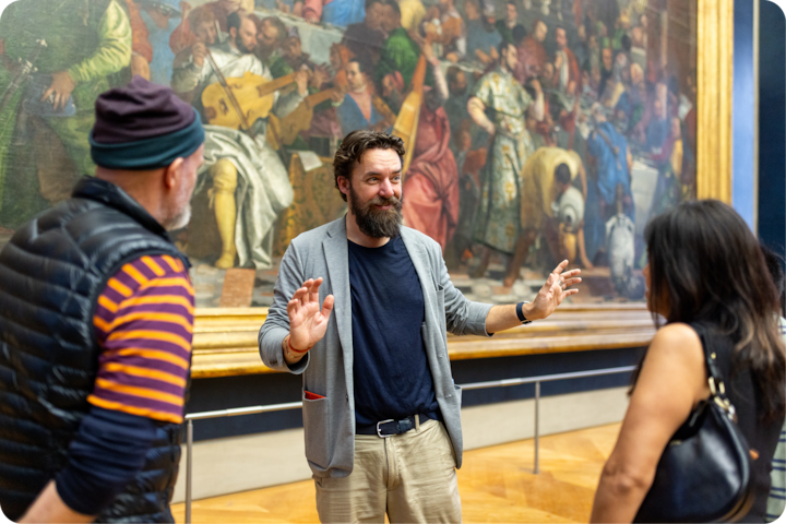 Un guide de musée fait des gestes devant une grande peinture colorée représentant une scène historique animée. Les visiteurs la contemplent attentivement.