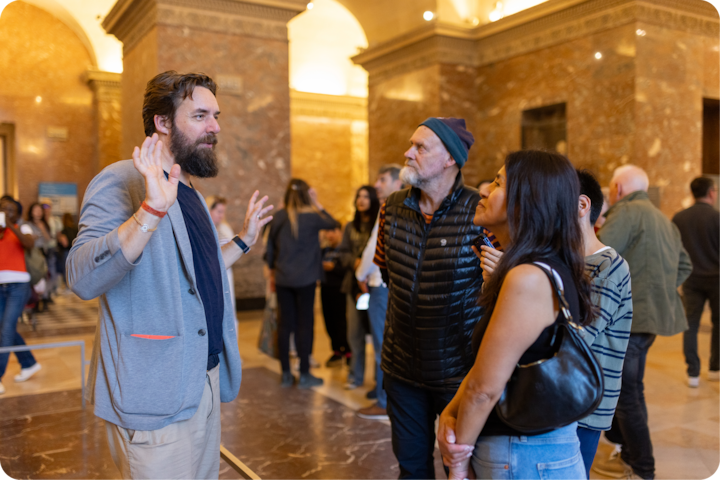 Dans un musée, des visiteurs attentifs se regroupent autour d'un guide dans une salle vaste à l'architecture ornementale et à l'éclairage chaleureux.