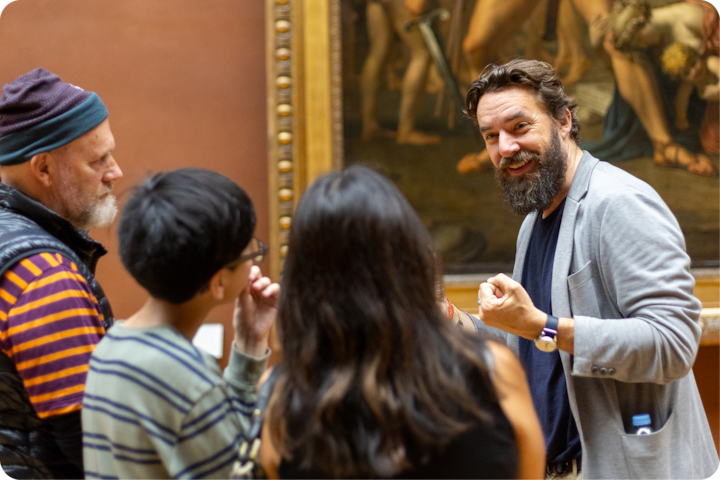 Des visiteurs de musée sont regroupés près d'une peinture classique, écoutant attentivement une personne qui fait des gestes en commentant l'œuvre.