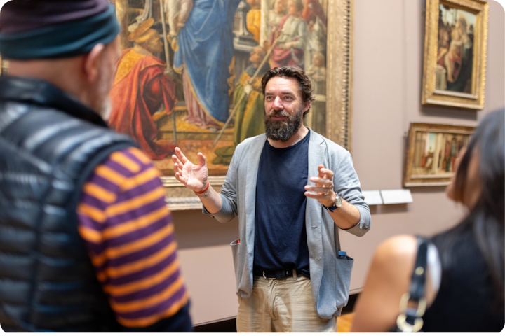 Une personne commente une œuvre d'art à un groupe de visiteurs du musée, réunis près de tableaux encadrés dans une galerie.