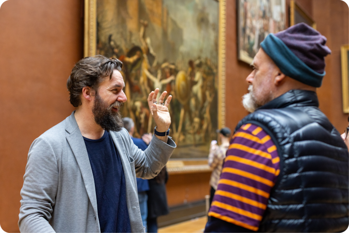 Un guide commente une œuvre d'art à un visiteur dans une galerie de musée présentant des tableaux historiques détaillés.