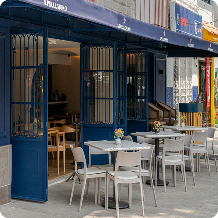 La terrasse d'un restaurant aux portes bleues et aux tables ornées de petits arrangements floraux, menant à un espace intérieur lumineux avec des chaises en bois.