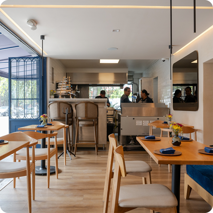 Restaurant convivial avec une salle unique, des sols en bois clair, une décoration épurée, des tables ornées de serviettes bleues et un bar donnant sur une cuisine ouverte.