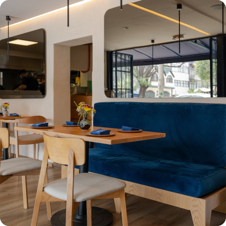 Intérieur d'une salle à manger avec une banquette bleue rembourrée, des chaises en bois et des tables dressées avec des serviettes bleues et des petits vases garnis de fleurs.