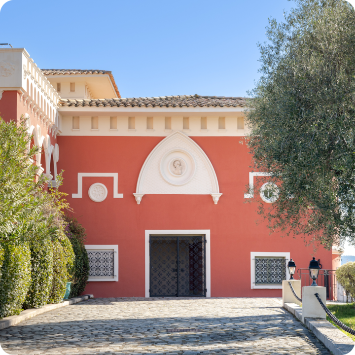 Vue extérieure d'un bâtiment rouge doté d'éléments blancs et d'un toit en tuiles, entouré d'un aménagement paysager luxuriant composé d'arbres et d'arbustes longeant une allée pavée.