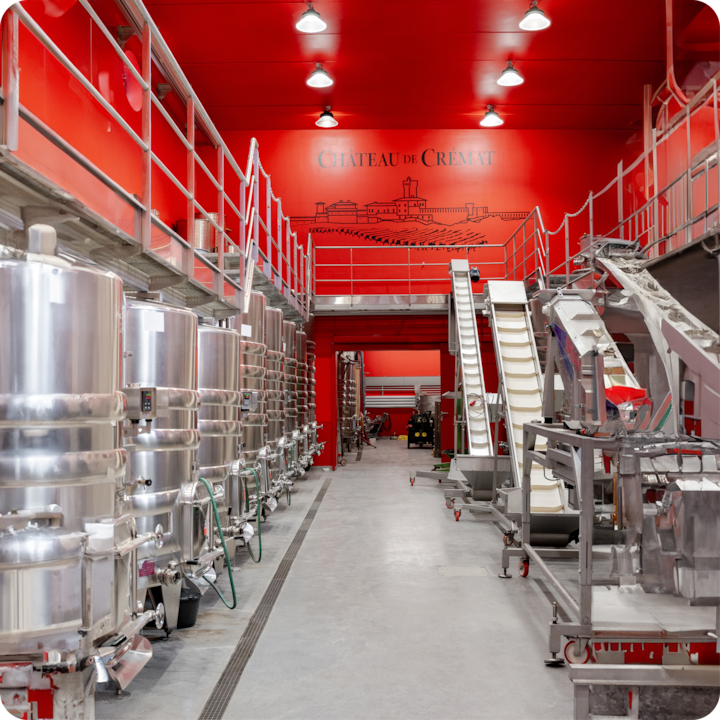 Une zone de production de vin lumineuse avec des cuves de fermentation en acier inoxydable, des machines et des murs rouges affichant une illustration « Château de Crémat ».