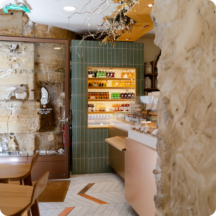 Un café lumineux aux murs carrelés de vert, doté d'une vitrine en verre pour les pâtisseries et d'une étagère garnie d'un assortiment de boissons, encadrées par des éléments de pierre rustique.
