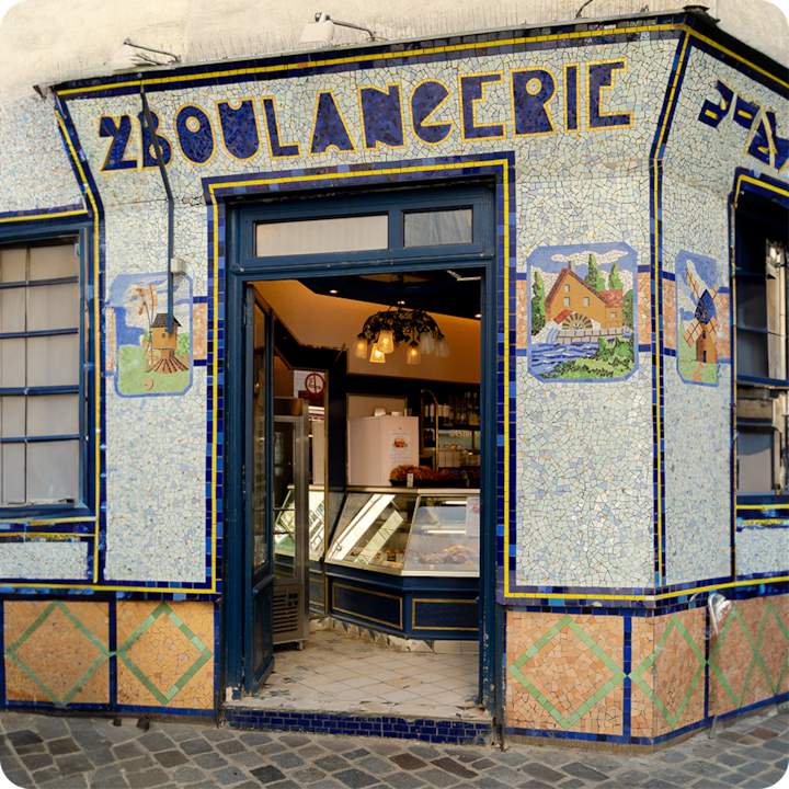 Une devanture en mosaïque avec l'inscription « Boulangerie » et une porte ouverte menant à l'intérieur de l'établissement, laissant apparaître des vitrines et des luminaires suspendus.