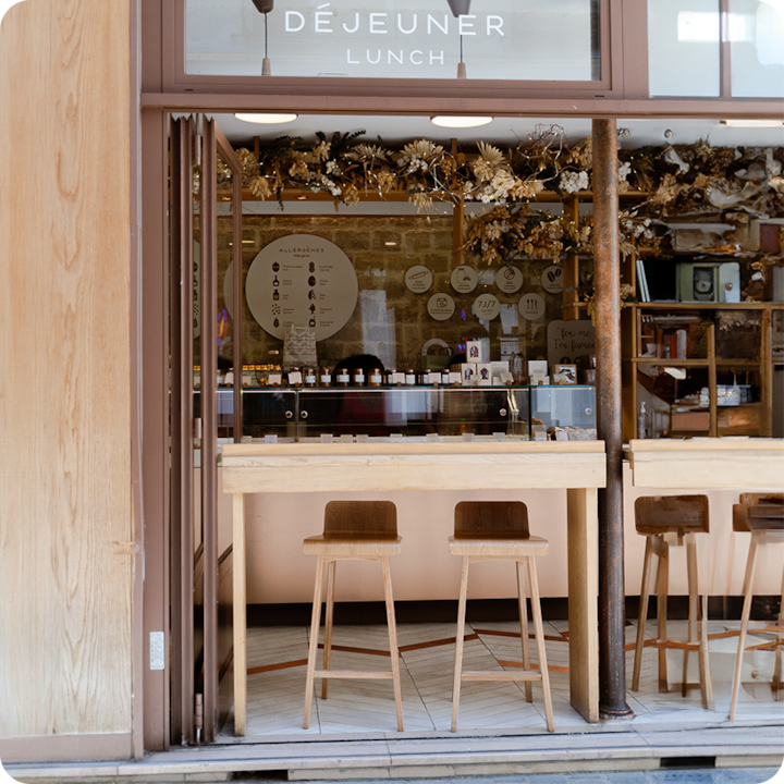 Vue extérieure d'un café avec des sièges en bois et un comptoir vitré, décoré de compositions florales séchées au-dessus de l'entrée.