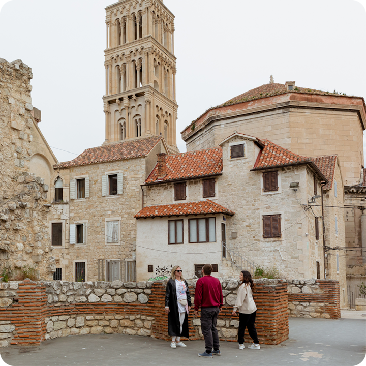 Un groupe de trois personnes se tient à l'extérieur devant des bâtiments historiques en pierre, dotés de toits en tuiles rouges et avec une haute tour en arrière-plan.