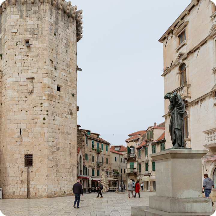 Statue en pierre sur une place historique entourée de bâtiments anciens en pierre aux volets verts et d'une grande tour.