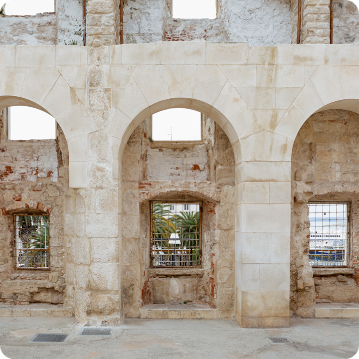 Un bâtiment historique en pierre avec arches et fenêtres, offrant une vue partielle sur des palmiers et une marina à travers des ouvertures grillagées.
