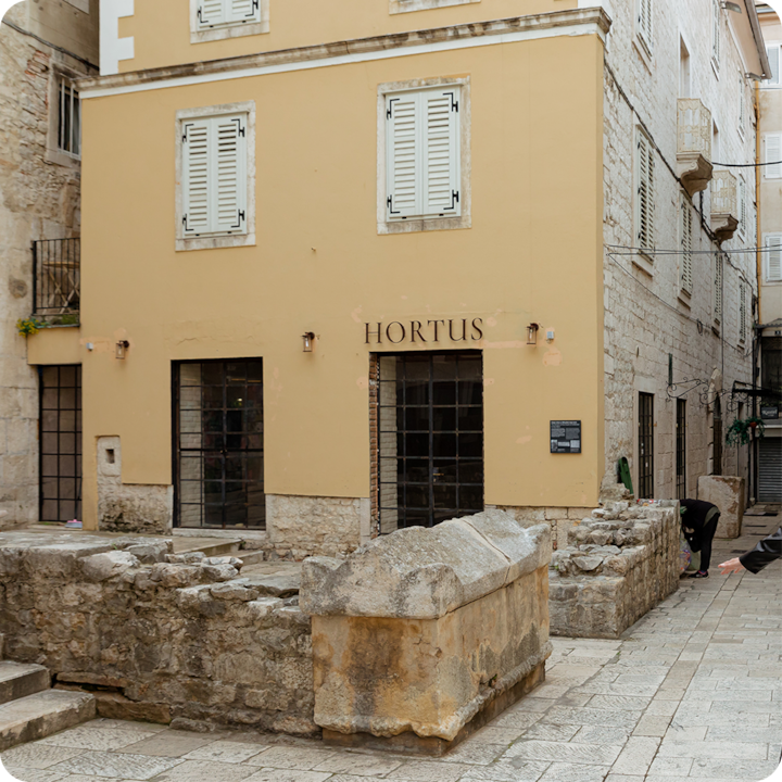 Un cadre extérieur historique mettant en scène un bâtiment beige entouré de ruines en pierre et d'un chemin pavé.