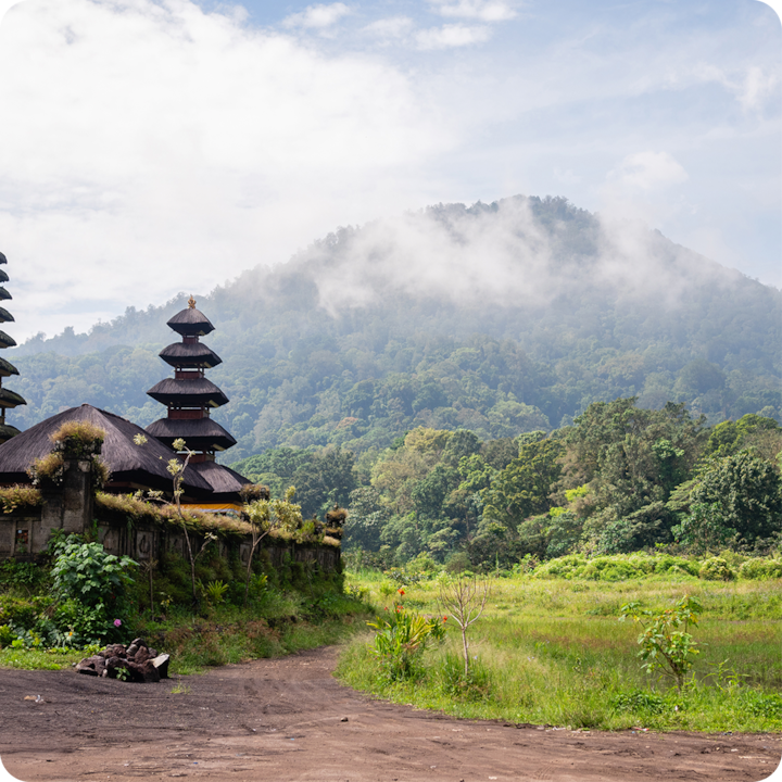 Un temple traditionnel à plusieurs niveaux entouré d'une végétation luxuriante, avec une montagne couverte de brume en arrière-plan sous un ciel partiellement nuageux.