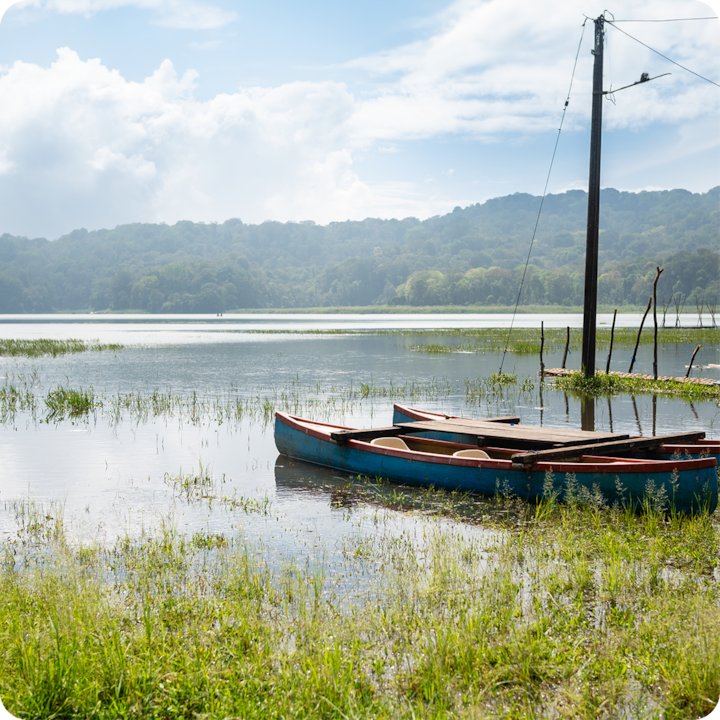Un lac paisible dans un cadre rural avec deux petits bateaux en bois ancrés près d'un rivage herbeux, entouré de collines verdoyantes sous un ciel nuageux.