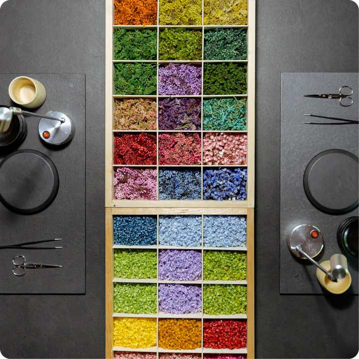 Un plan de travail noir présentant une boîte en bois remplie d'un assortiment de fleurs séchées colorées, des ciseaux, des pincettes et de petits récipients.