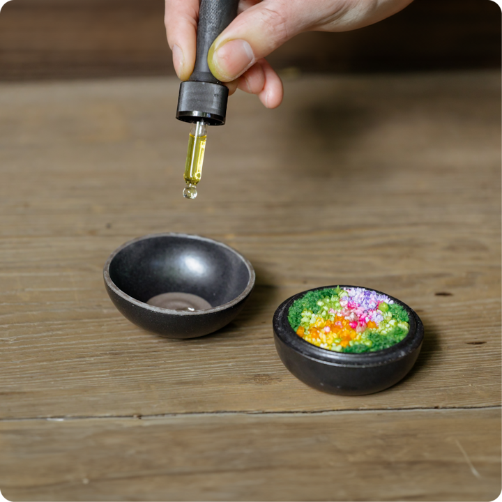 Une main utilise un compte-gouttes pour verser un liquide jaune dans un bol noir, placé à côté d'un bol de fleurs séchées colorées et de feuillages, le tout sur une table en bois rustique.