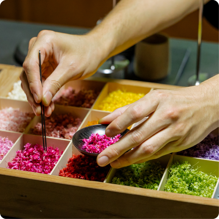 Gros plan sur des mains qui disposent des pétales de fleurs séchées aux couleurs vives dans une boîte en bois à compartiments, mettant en valeur une variété de matériaux colorés.