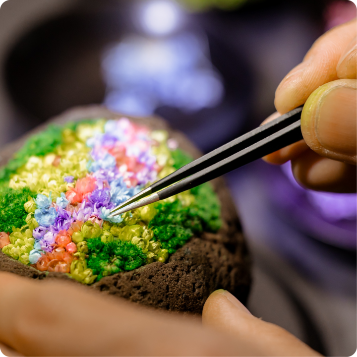 Des mains positionnent délicatement à l'aide de pincettes de minuscules fleurs roses, violettes, bleues et jaunes sur une base verte et brune texturée.