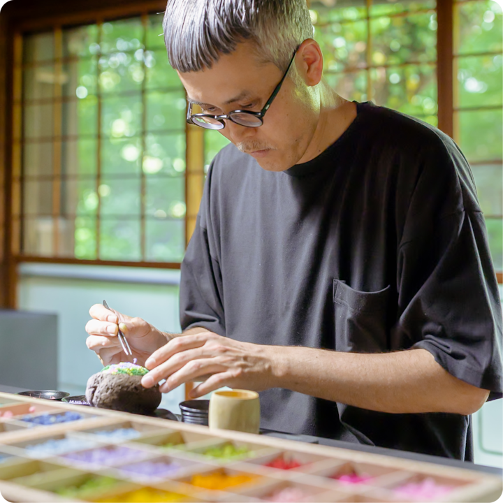 Une personne dispose soigneusement des matériaux colorés sur un petit objet, devant un plateau en bois garni de poudres colorées. De grandes fenêtres laissent entrevoir un environnement vert apaisant.