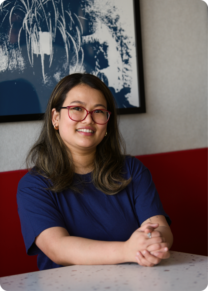 Une personne est assise, les mains croisées sur une table mouchetée. Un mur rouge et une peinture abstraite en bleu et blanc sont visibles en arrière-plan.