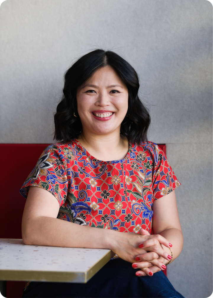 Une personne assise portant un haut rouge vif avec des motifs floraux et géométriques détaillés. Ses mains reposent sur une table blanche à bord doré.