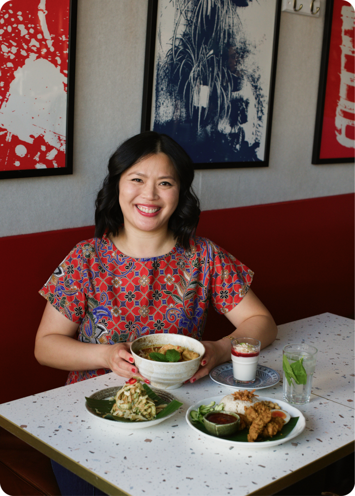 Un assortiment de plats inspirés de l'Asie du Sud-Est est disposé sur une table blanche mouchetée. On y trouve du poulet frit, du riz, de la soupe, de la salade et un dessert en couches. Des œuvres d'art abstraites sont accrochées au mur en arrière-plan.