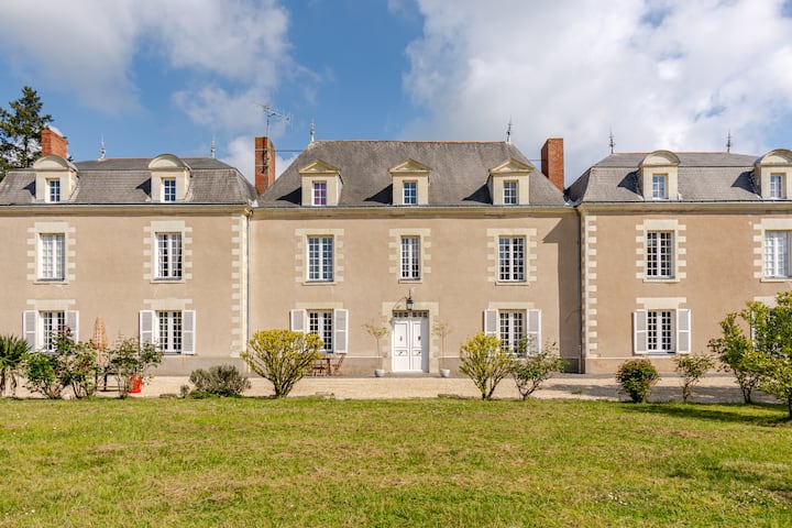 Un imposant manoir beige avec des toits ornés, une architecture symétrique et des buissons paysagers, photographié par une journée ensoleillée avec quelques nuages dans le ciel.