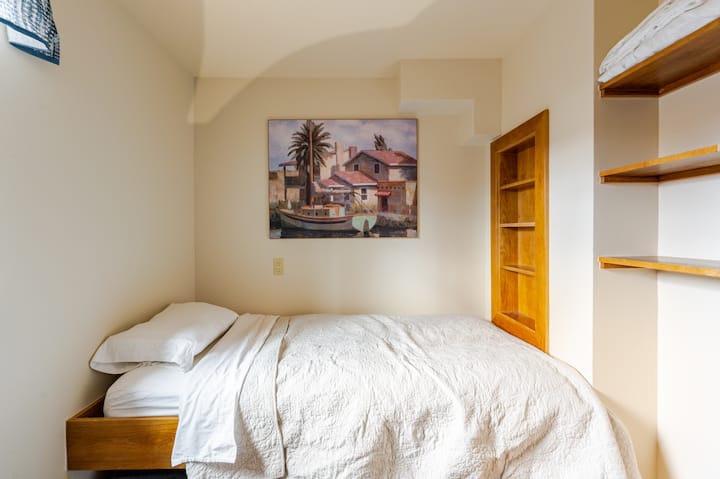 Une petite chambre baignée de lumière naturelle, avec un lit recouvert de draps blancs, des éléments en bois et un tableau coloré représentant un village côtier accroché au mur.