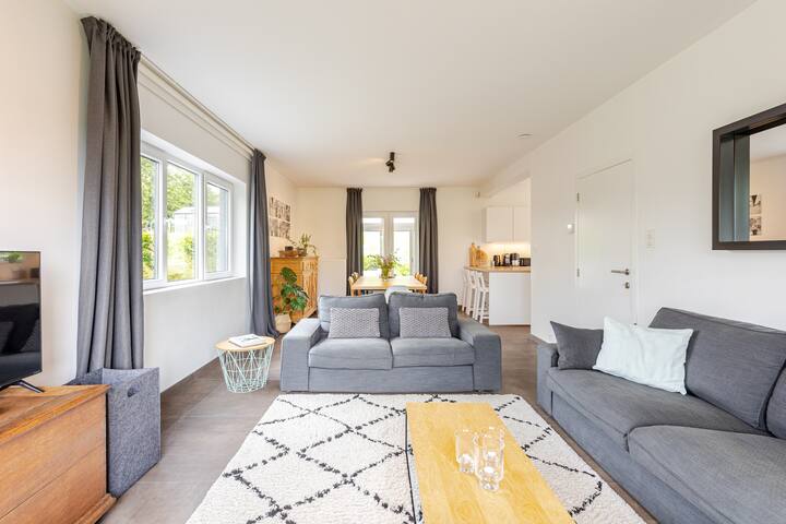 Un salon spacieux et ouvert avec un mobilier contemporain, de la lumière naturelle provenant de grandes fenêtres et une cuisine attenante en arrière-plan.