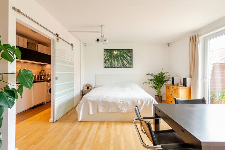 Un studio contemporain avec un lit blanc, des plantes en pot, des meubles en bois et une table à manger, ainsi qu'une porte vitrée coulissante menant à un coin cuisine.