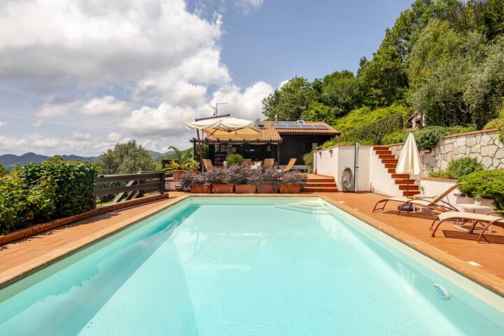 Une piscine extérieure paisible avec un sol en terre cuite, un patio ombragé avec des sièges et une végétation luxuriante, nichée au milieu de collines verdoyantes et de forêts.