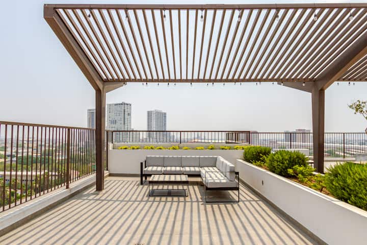 Un espace lumineux sur un toit avec une pergola à lattes en bois, des sièges, des plantes luxuriantes et une vue sur l'horizon urbain sous un ciel dégagé.