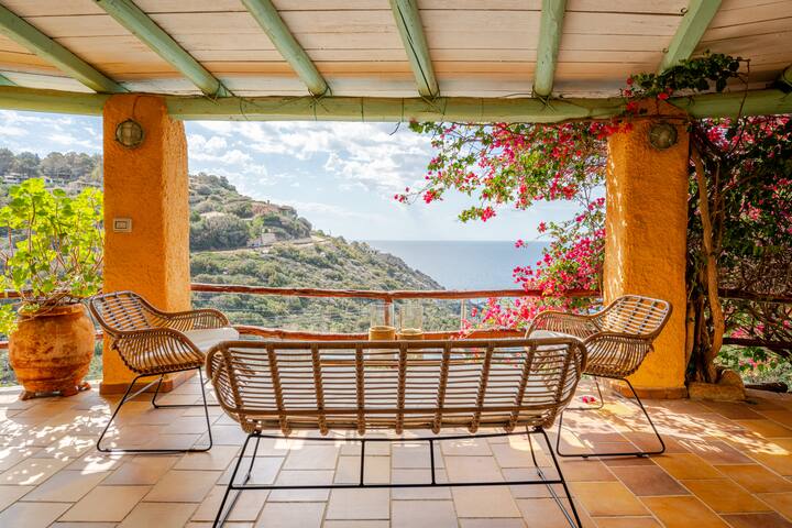 Véranda rustique avec des chaises en osier, des bougainvilliers en fleurs et une vue panoramique sur le littoral et la végétation.