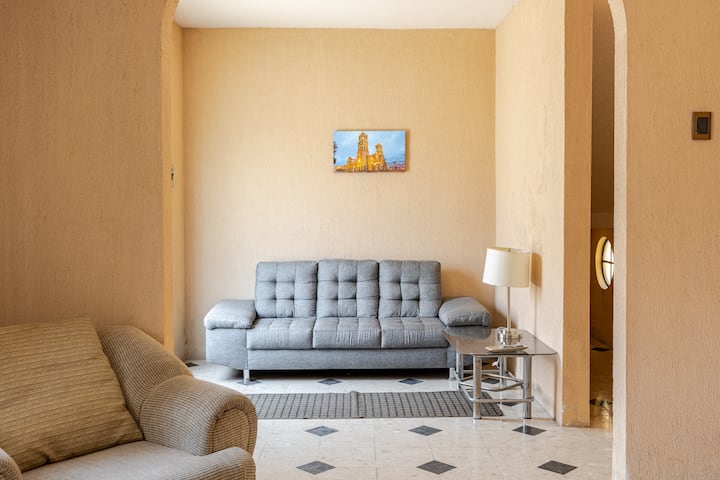 Un salon minimaliste composé d'un canapé gris, d'un fauteuil beige et d'une table d'appoint avec une lampe, encadré par des arcades. Un tableau représentant une cathédrale décore le mur au-dessus du canapé.