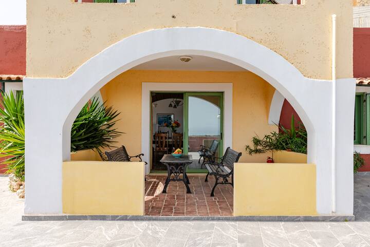 Un bâtiment de style méditerranéen avec une arche blanche, des murs jaunes, des plantes et un patio équipé de chaises en métal et d'une table, menant à un espace repas à l'intérieur.
