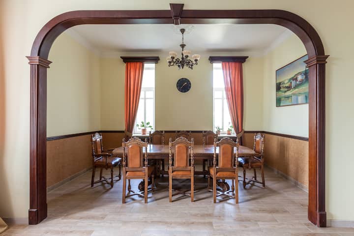 Un espace repas formel avec une longue table en bois et des chaises, éclairé par la lumière naturelle de deux grandes fenêtres ornées de rideaux orange. Un lustre central, une horloge murale et des tableaux composent un décor classique, encadré par une arche en bois.