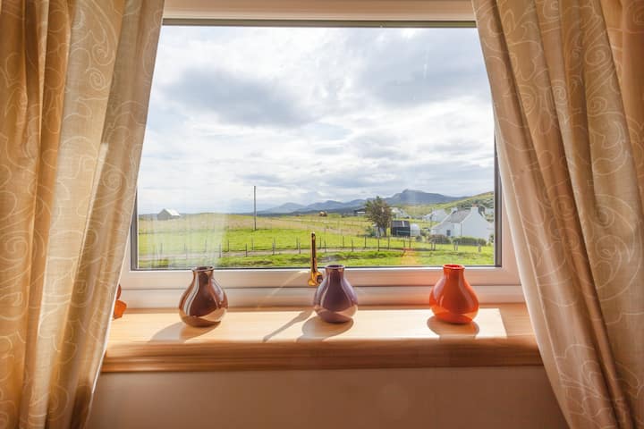 Vue depuis une fenêtre sur un paysage rural avec des pâturages verdoyants, des collines vallonnées et quelques maisons sous un ciel nuageux. Des rideaux beiges et des vases colorés agrémentent le rebord de la fenêtre.