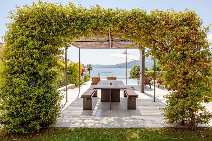 Un coin repas extérieur encadré par un treillis en bois couvert de verdure luxuriante, surplombant une piscine et un lac pittoresque avec des montagnes au loin.