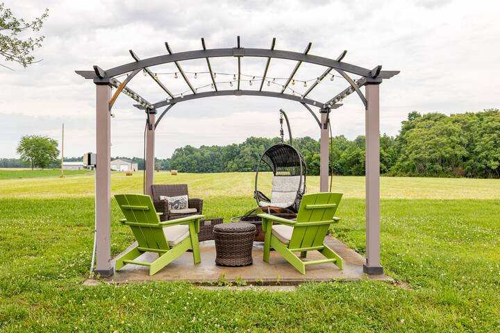 Une pergola confortable avec des chaises vertes, du mobilier en osier et un fauteuil suspendu dans un vaste champ, offrant une expérience de détente en plein air.